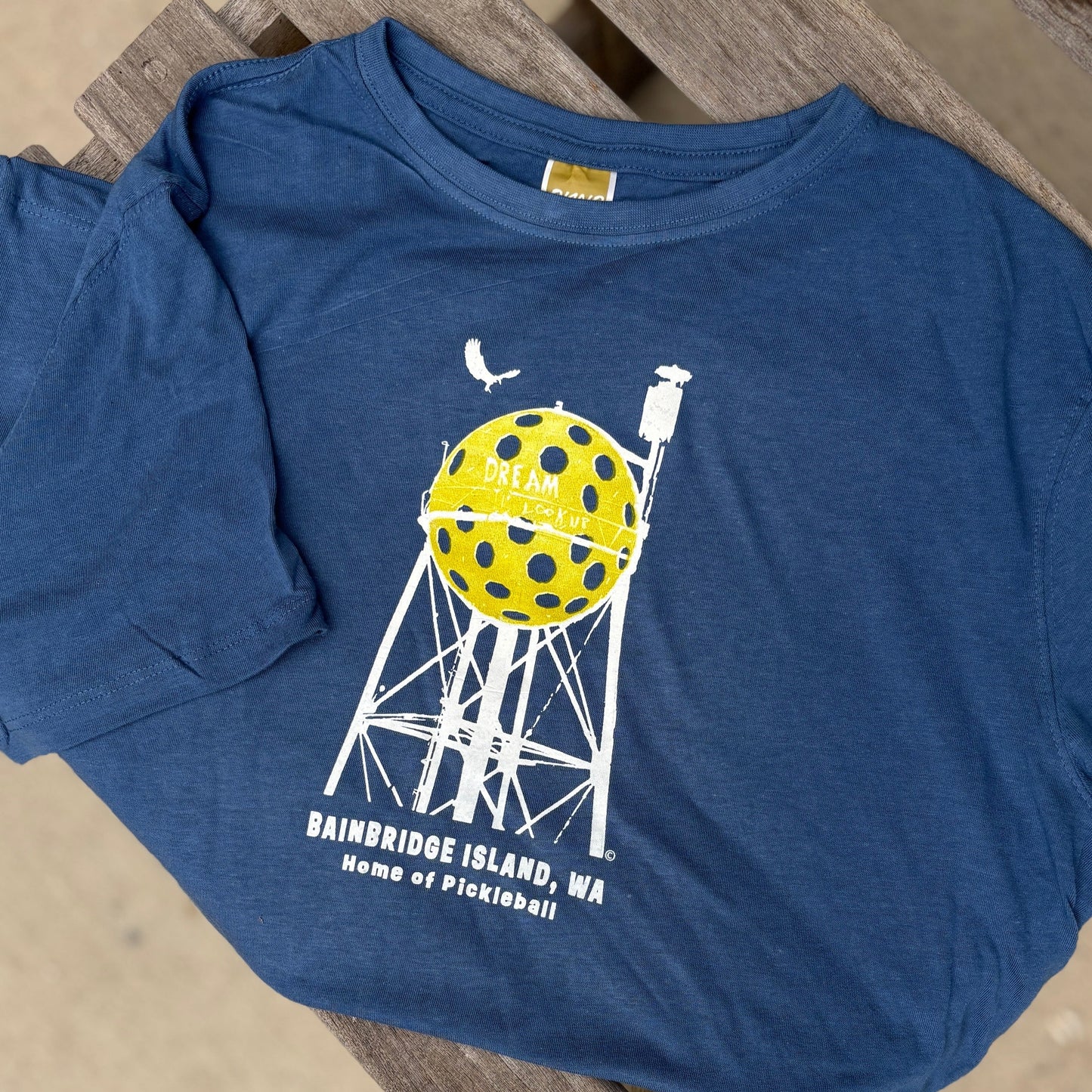 Blue t-shirt with a yellow and white graphic  pickleball tower on a wooden surface